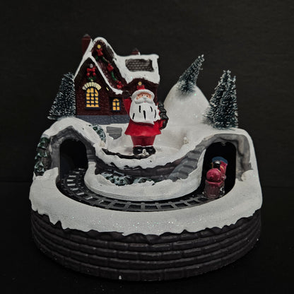 A rotating Christmas village house with a snowman, Santa Claus, and a small cabin in a snowy setting.