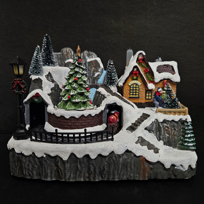 A holiday-themed decoration featuring a snowy scene with a Christmas tree, house, and figurines of people enjoying the snow.